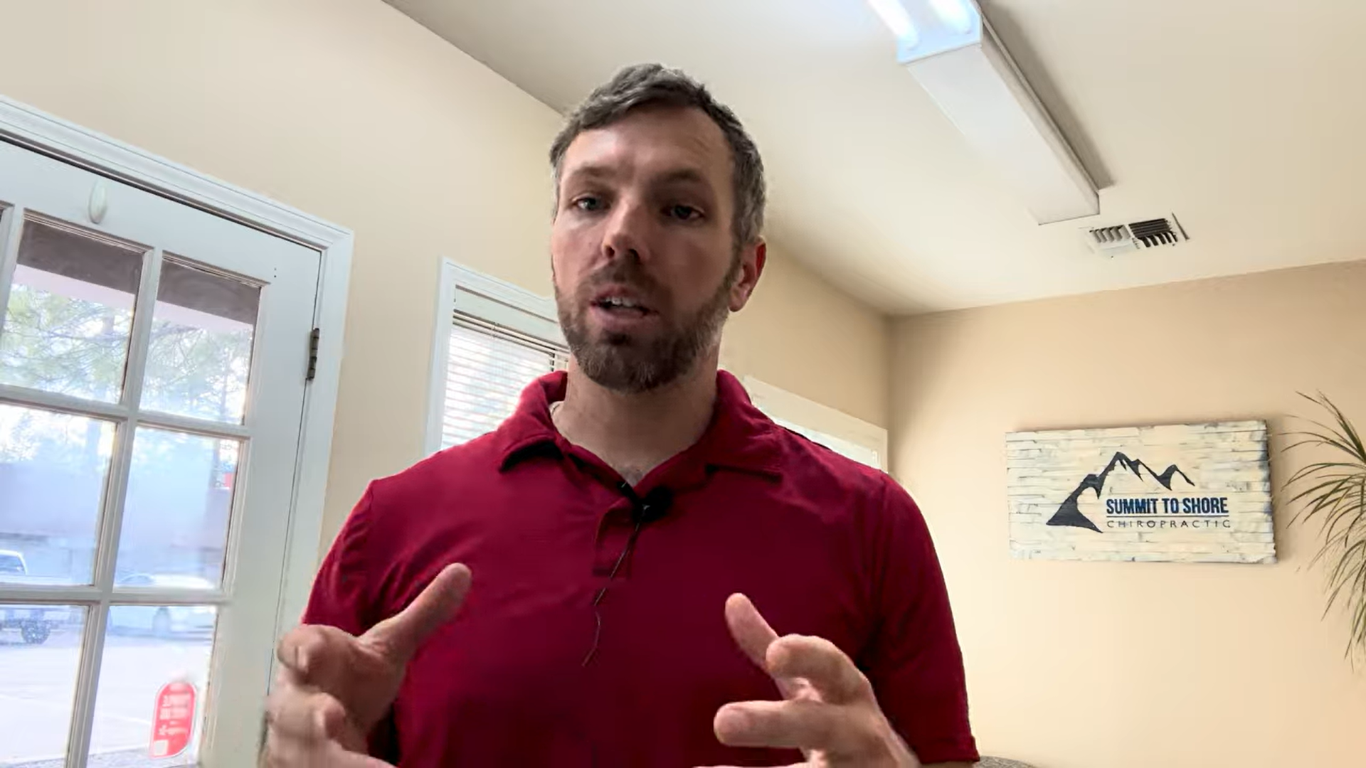 Man in a red polo speaking to camera in a bright clinic, with a 'Summit to Shore Chiropractic' sign on the wall behind him.