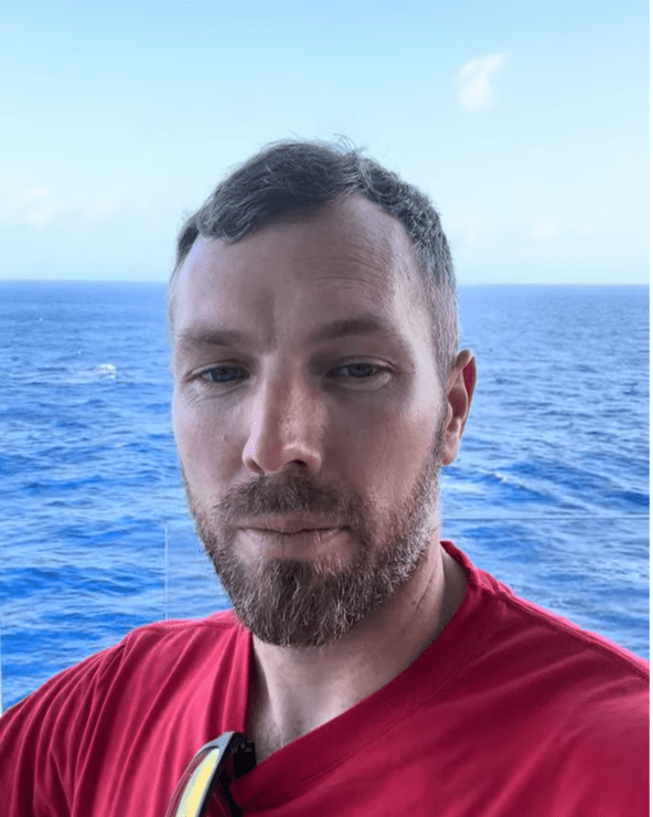 Screenshot (34) Close-up portrait of a man with short brown hair and a beard, wearing a red shirt, with the blue ocean and sky in the background.