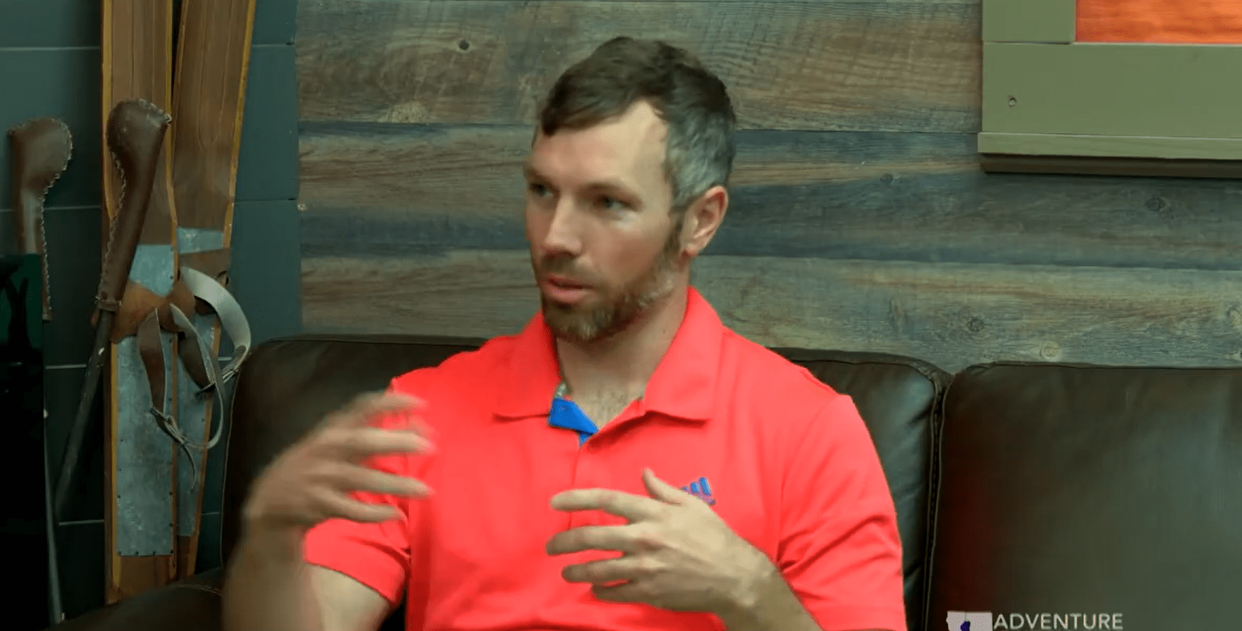 Screenshot (36) Man with short brown hair and beard wearing a coral polo shirt sits on a brown leather couch, gesturing while talking against a wooden wall.